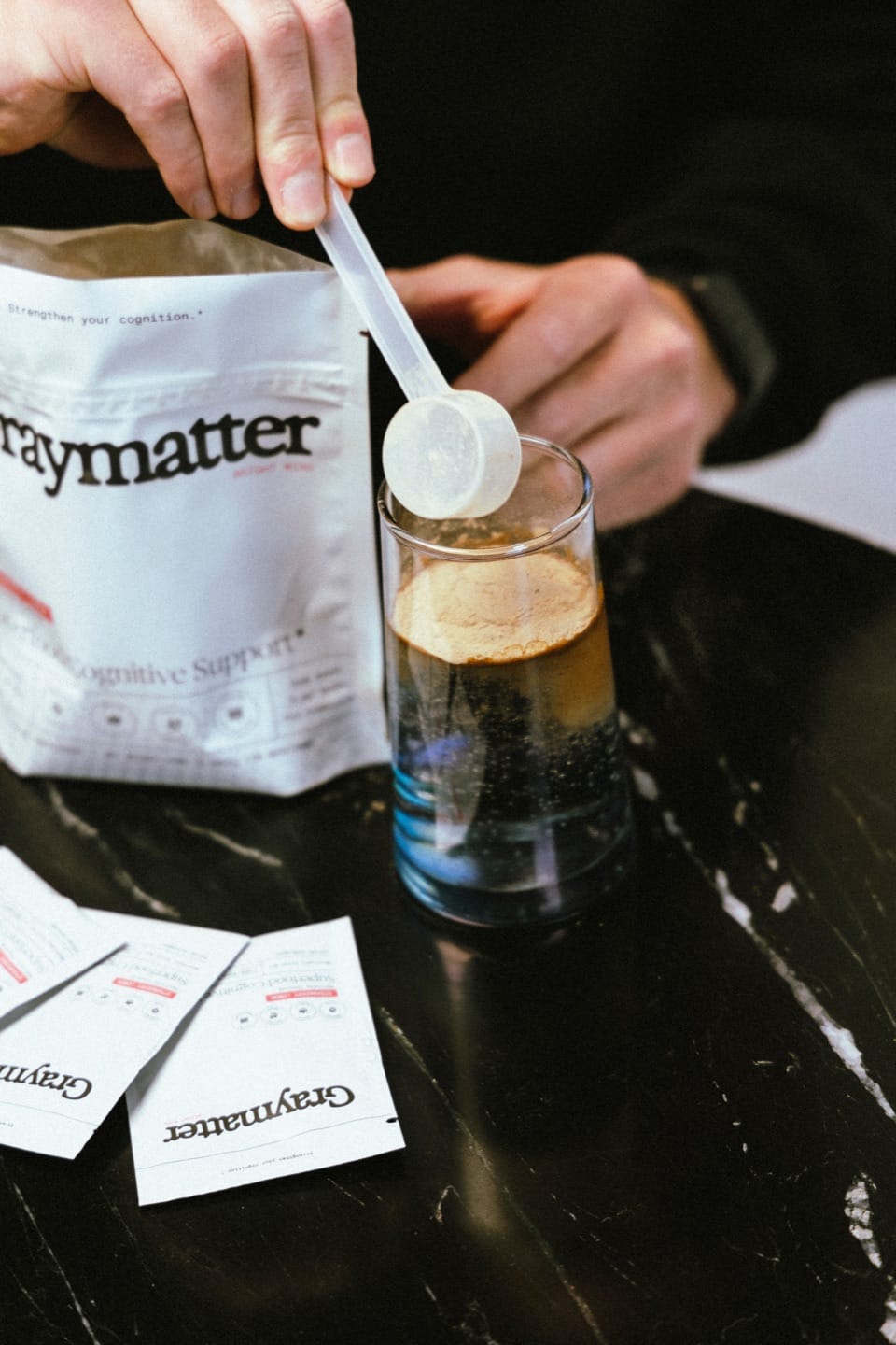 A scoop of Graymatter powder being added to a glass of water next to the Graymatter pouch and travel packs